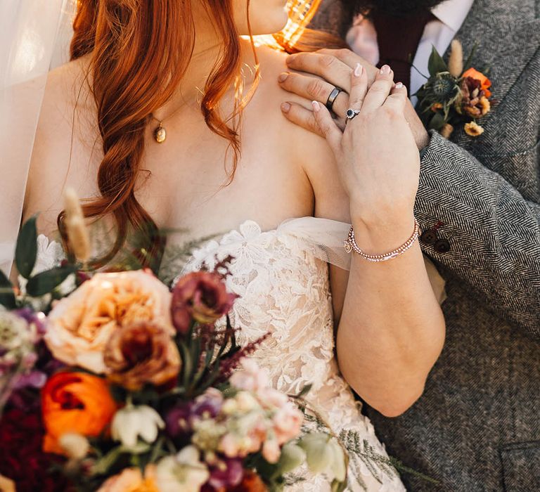 Bearded groom in a grey suit stands closely with the bride who wears gold wedding jewellery 