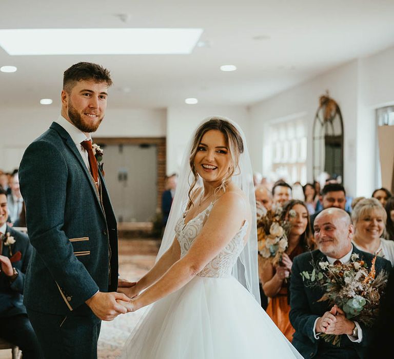 Bride and groom smile happily as they hold hands for their ceremony as both of their fathers smile at each other 