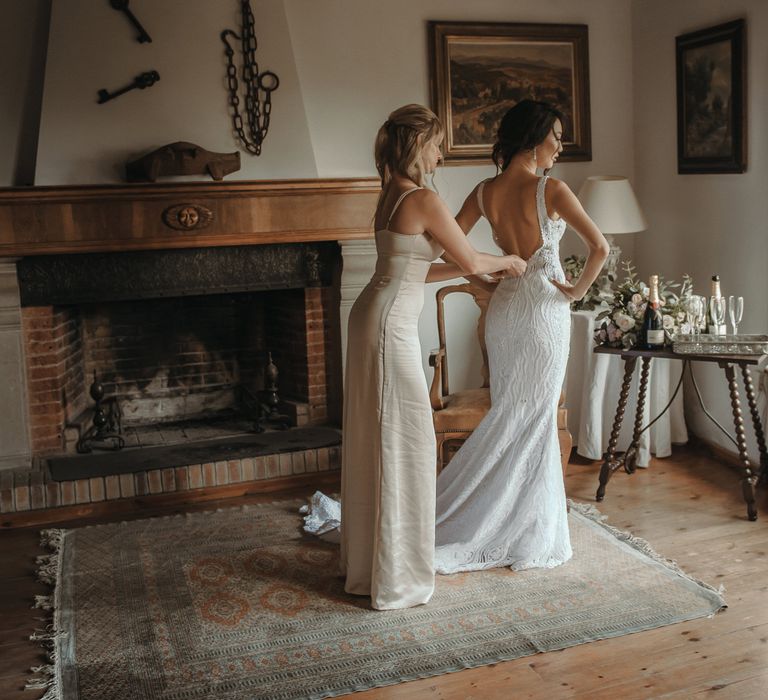 Bridesmaid wearing champagne coloured bridesmaid dress does up the brides wedding dress in front of rustic fireplace 