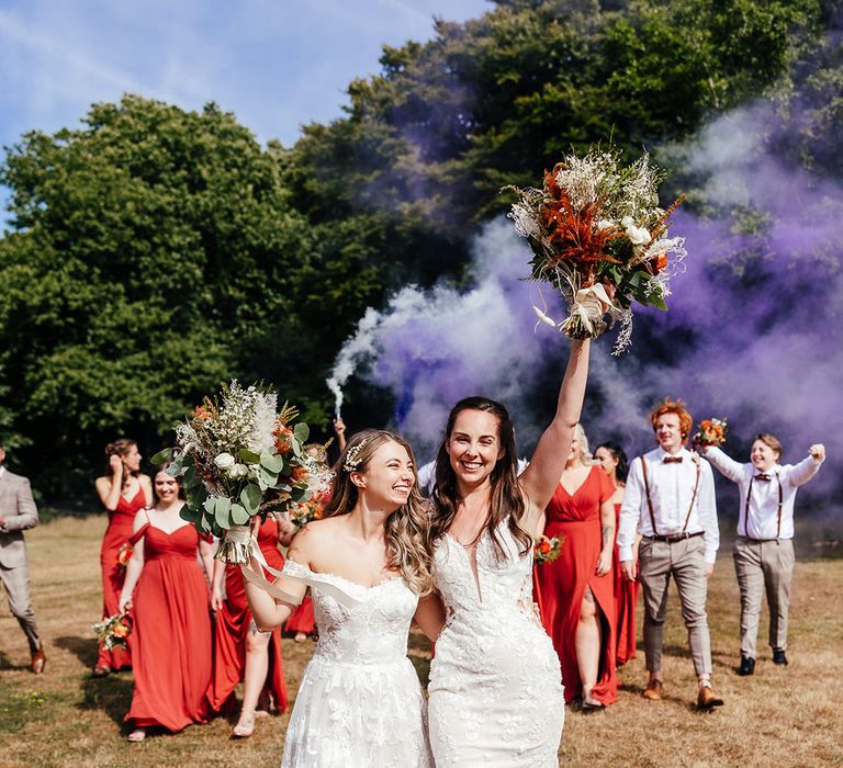 Brides raise their bouquets in the air with bridesmaids in orange dresses and grooms in bow ties, braces and shirts letting off purple smoke bombs