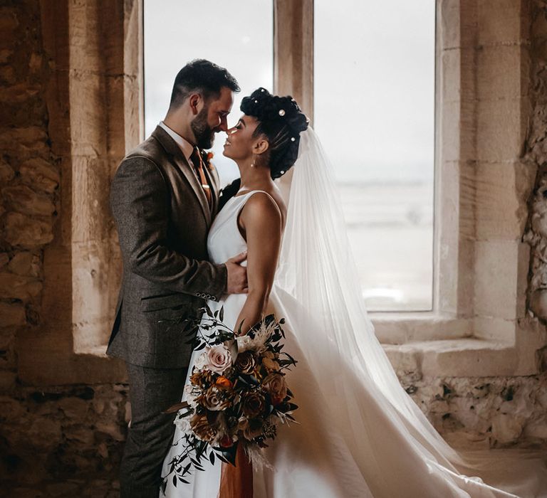 Bride wears her floor length veil to the back of her braided hair whilst kissing embracing her groom in three piece suit in front of bright window