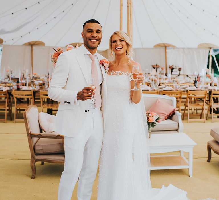 Bride in off the shoulder wedding dress with a glass of champagne and groom in a white wedding suit and pink tie for their marquee wedding