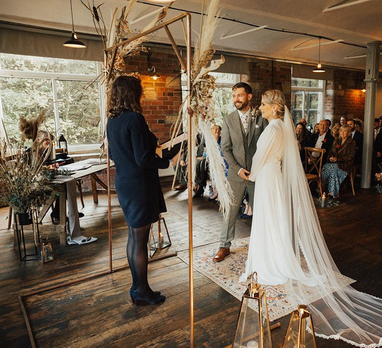 Bride in Reformation wedding dress & groom stand as brass arch complete with dried floral arrangements and 