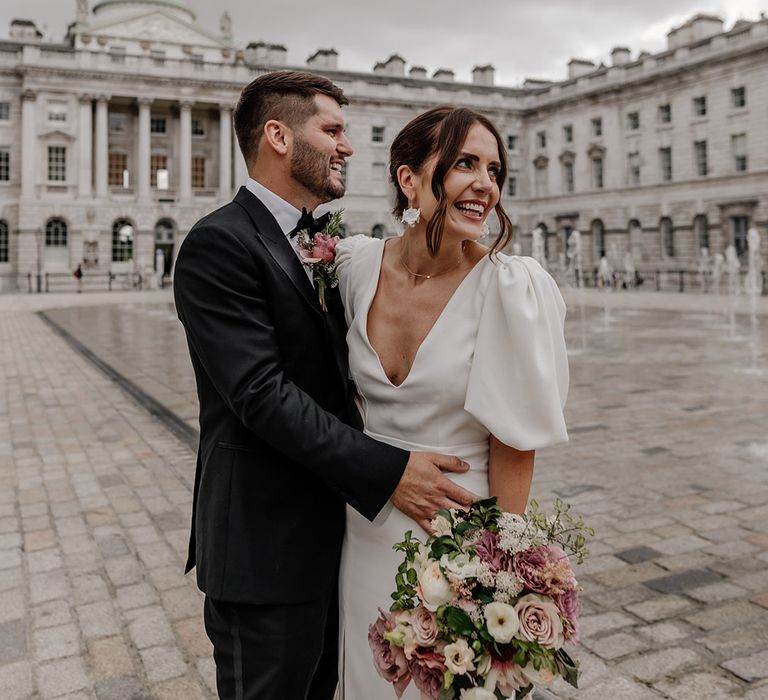 Bride in front slit v neck wedding dress with puff sleeves embracing groom in dark tux at Somerset House wedding