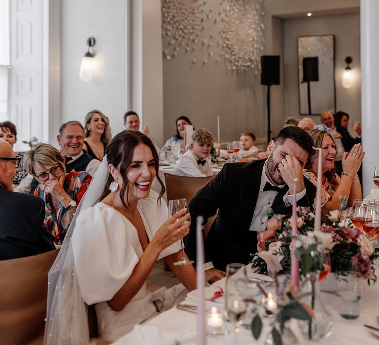 Bride, groom and wedding guests sitting and laughing at wedding reception underneath large avant garde bauble chandelier