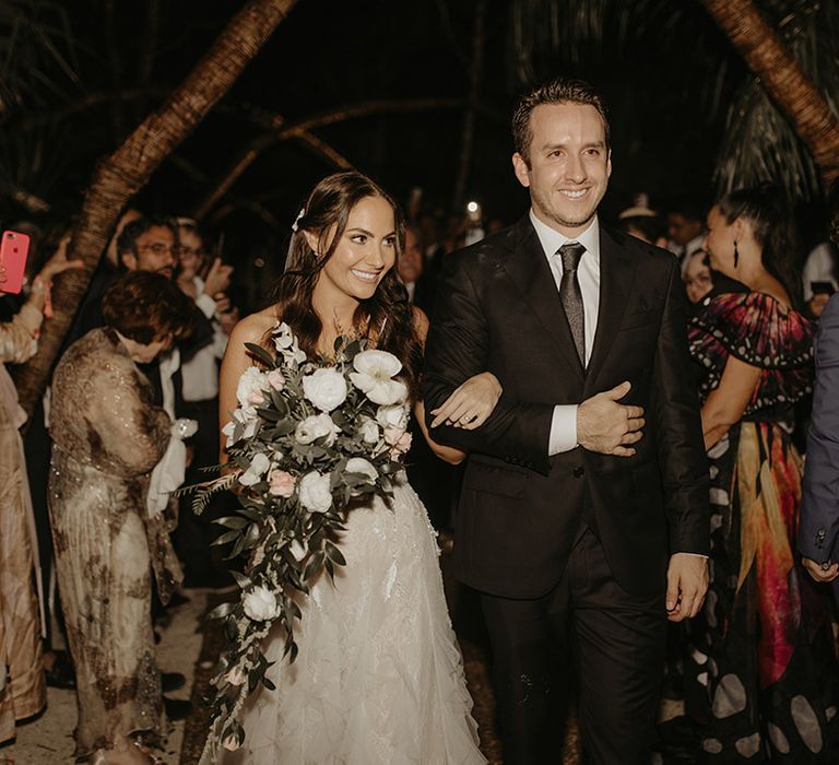 Bride holding white floral bouquet with green foliage walks arm in arm with groom 