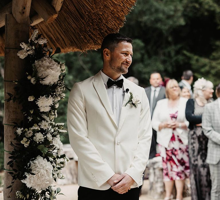 The groom in a white suit jacket and black tie with a white rose buttonhole stands smiling as he sees the bride walking to him 