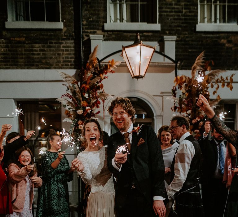 Bride in long sleeve lace vintage wedding dress and closed toe satin heels and groom in dark suit with orange tie and autumnal boutonniere doing sparkler exit from autumnal intimate London wedding