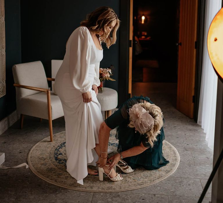 Bride steps into her white block heel wedding shoes with pearls as her mum helps fasten them 