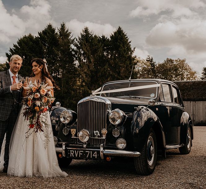 Shiny black vintage wedding car decorated with white ribbon with the bride in a lace and tulle wedding dress and groom in a checkered grey suit standing in front of it 