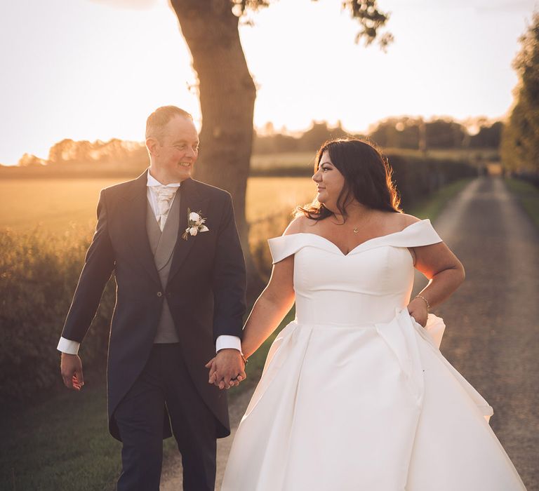 Rustic Stratton Court Barn Bicester wedding venue with the bride in an off the shoulder dress with the groom in a morning suit walking during sunset