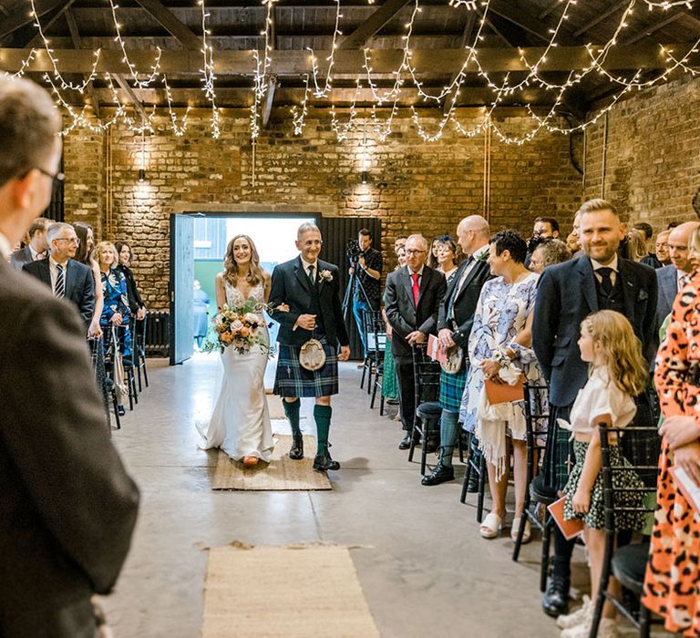 Fairy lights hang from ceiling as bride walks down the aisle during industrial styled wedding ceremony 