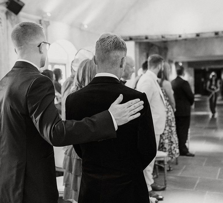 Best man puts his arm around the groom to support him as the bride begins her walk down the aisle 