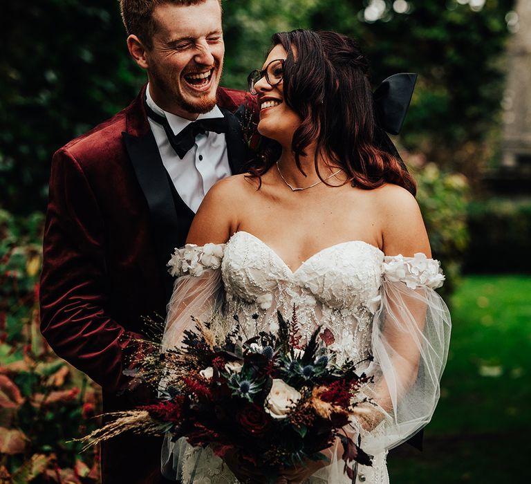 The bride wears an off the shoulder sheer puff sleeve wedding dress and big black hair bow accessory with the groom in a burgundy suit 