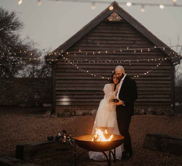 Bride in square neck wedding dress with sheer puff sleeves embracing groom in in classic black tuxedo with black bowtie standing by the fire pit 