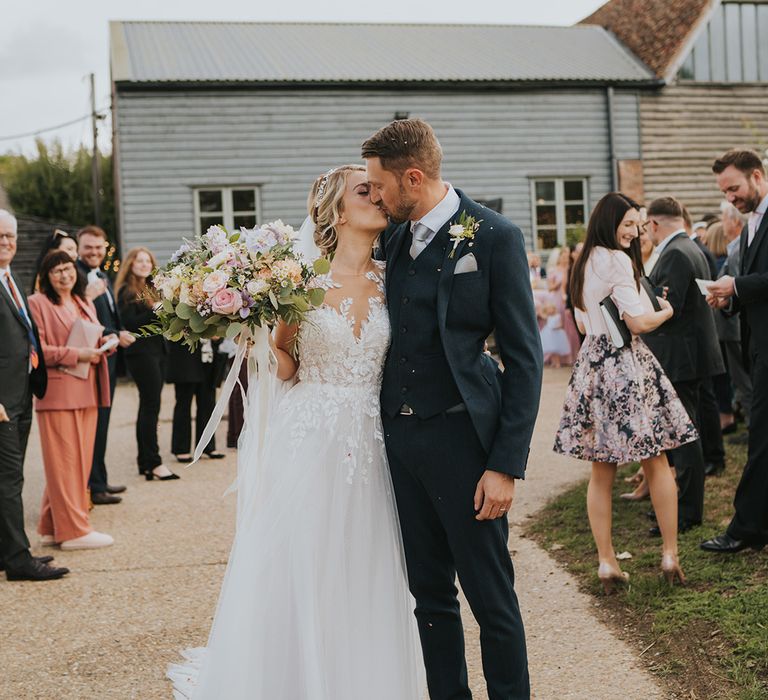 The bride and groom share a kiss after their confetti exit with the bride in an illusion lace wedding dress and navy suit 