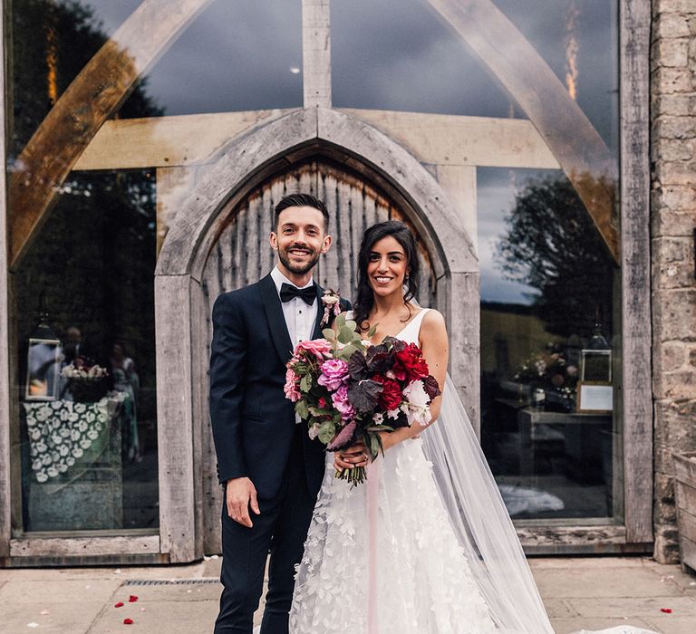 Bride in flower appliqué wedding dress and cathedral length veil posing with the groom in a classic black and navy tuxedo 