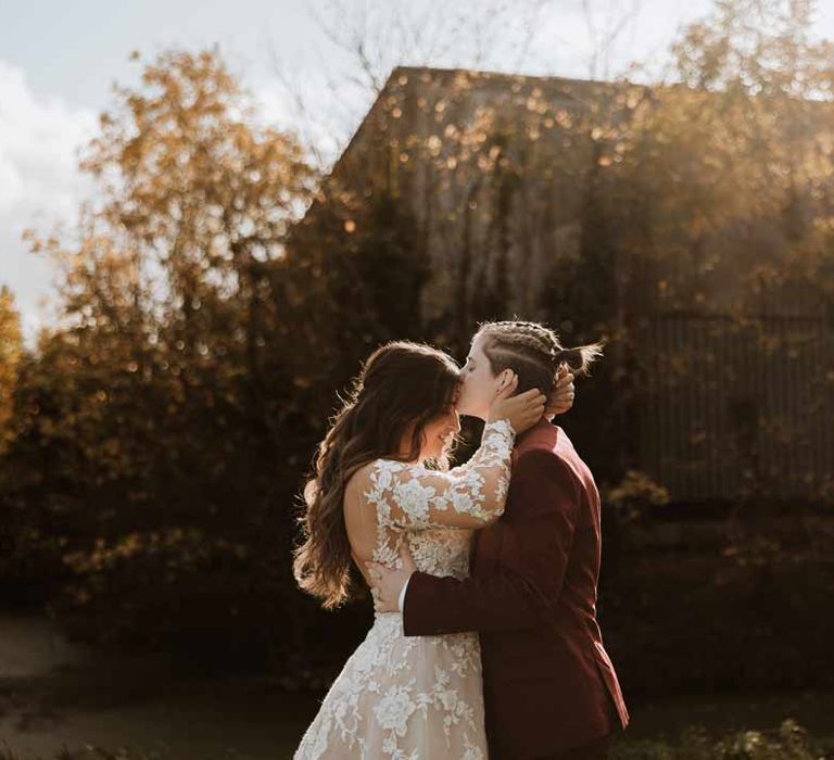 Bride wearing maroon bridal suit with grey waistcoat, dusky pink tie and autumnal dried flower boutonniere kissing bride on the forehead wearing long sleeve lace illusion sleeve wedding dress with overlay 