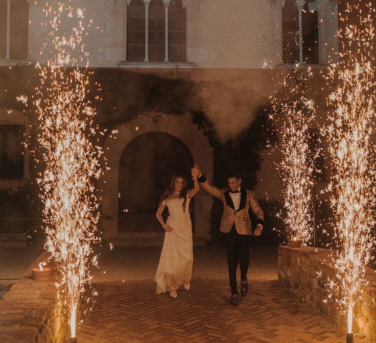 Bride in off the shoulder reception dress with groom in gold blazer with black lining, black bowtie and black suit trousers by large standing firework display  