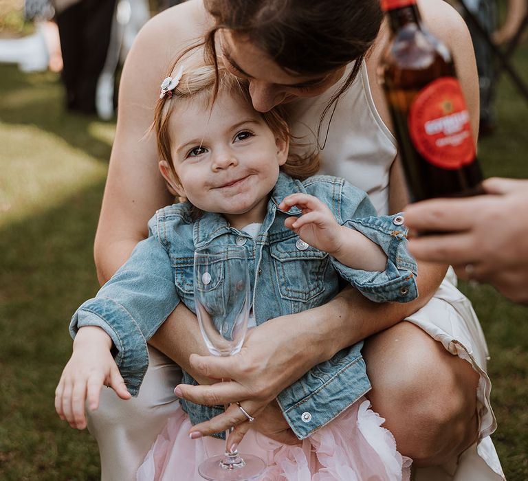 Little wedding guest in a pink tulle skirt and light blue denim jacket smiling with a bridesmaid in cream satin dress 