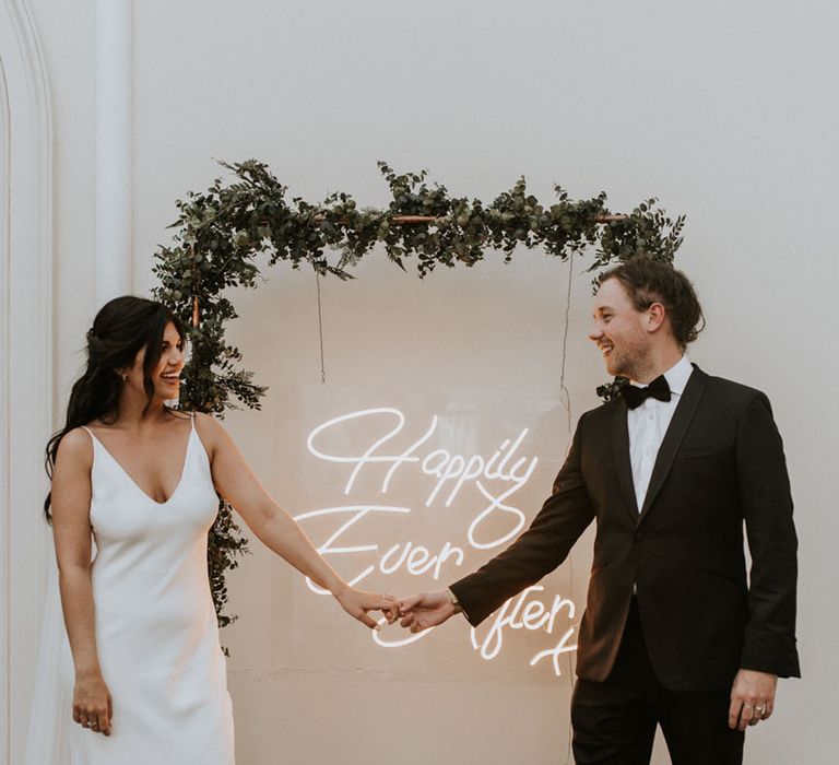 Bride in plunging wedding dress with the groom in front of a white neon wedding sign with foliage decor 