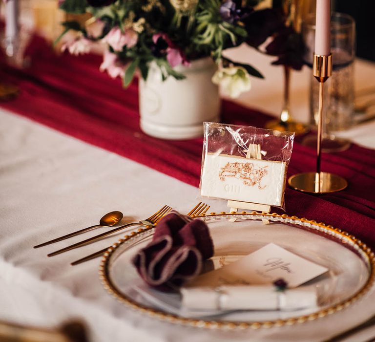 Iced wedding biscuits with gold cutlery, gold charger plate, burgundy napkin and table runner with purple floral arrangement and taper candle