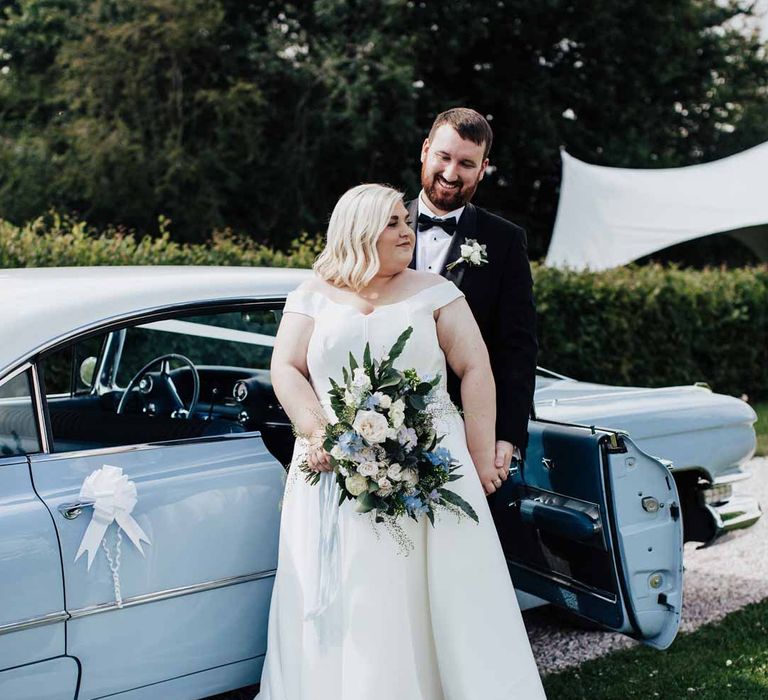 Groom in classic black grooms three piece tuxedo with white rose boutonniere and black bowtie embracing bride in satin off the shoulder wedding dress holding white and blue eucalyptus, foliage, garden rose, gardenia, light blue delphinium and dried flower bridal bouquet tied with dusky blue ribbon standing by classic baby blue wedding car