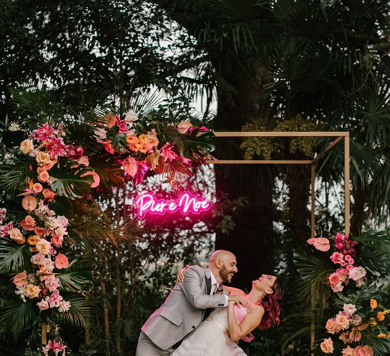 groom in a grey wedding suit leaning his bride over for a kiss in front of their gold frame altar decorated with tropical pink wedding flowers and a personalised neon pink wedding sign