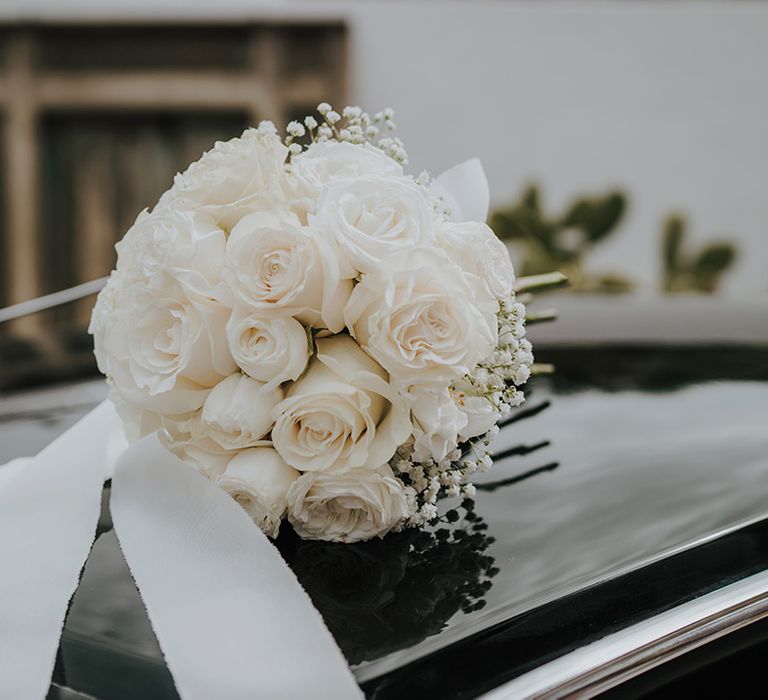 white rose and gypsophila wedding bouquet tied with ribbon sitting on a black car bonnet