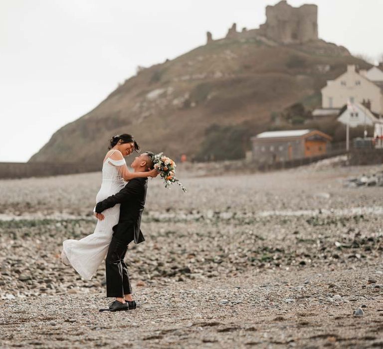 Groom in classic black tux picking up and spinning bride in white beaded and sequined slip wedding dress for coastal wedding photoshoot 