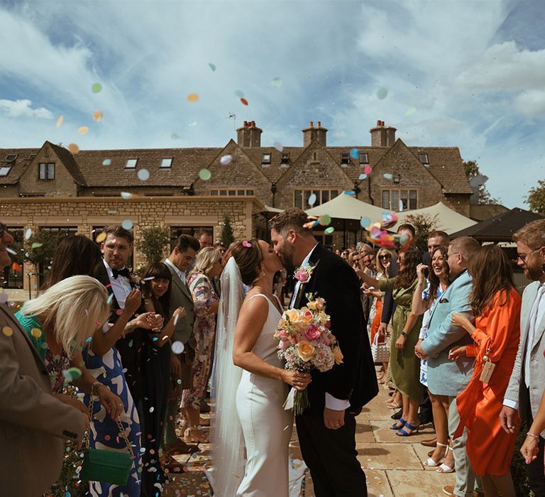 The bride and groom kiss at their confetti exit from their outdoor ceremony with guests throwing confetti 