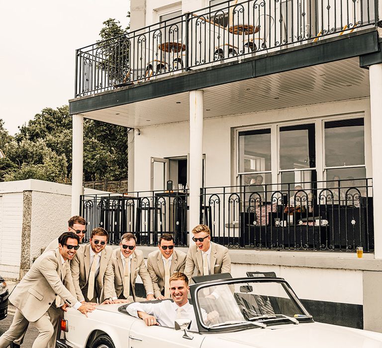 groomsmen in beige suits pushing the groom in a light grey suit in his white covetable wedding car