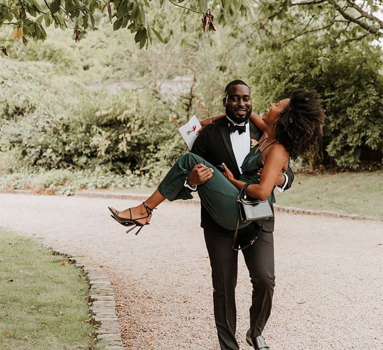 Groomsman in black tuxedo carrying a wedding guest in green dress 