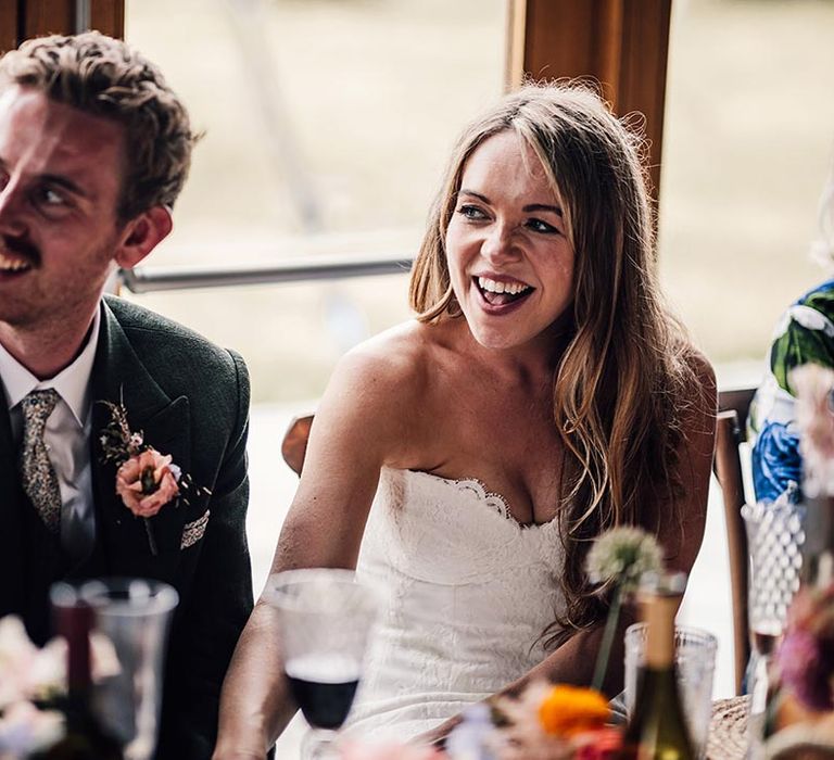 Groom in dark green suit who floral tie sitting with bride in strapless gown at the wedding reception 