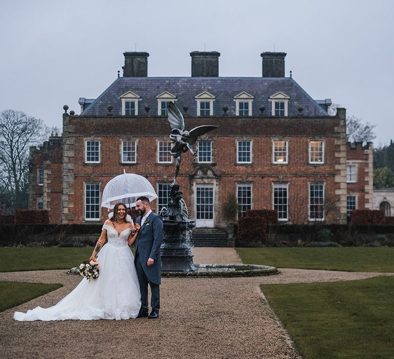 The bride and groom walk underneath and umbrella on rainy wedding day at country house wedding venue 