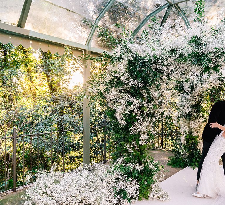 The bride and groom kiss under the white gypsophila chuppah at the glasshouse 