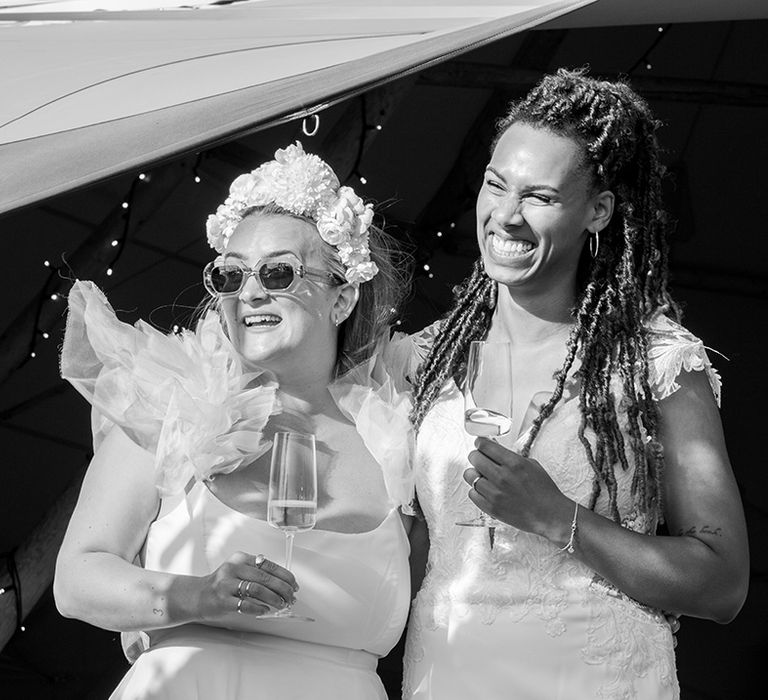 Two brides holding drinks smile as they listen to the wedding speeches 