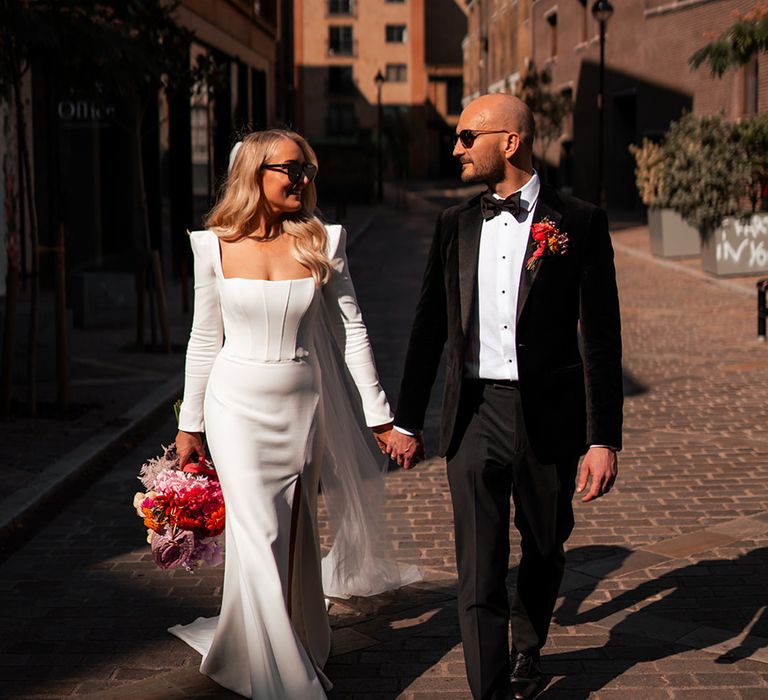 Bride in shoulder pad Suzanne Neville wedding dress with sunglasses walking hand in hand with groom in black tux at London wedding