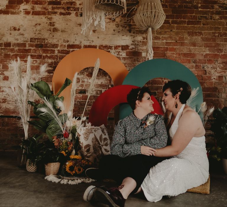 Brides in a jumpsuit and patterned shirt sitting by a colourful altar styled with dried flowers 