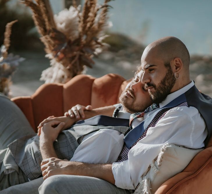 The grooms lie together on velvet scalloped seating at their outdoor wedding reception