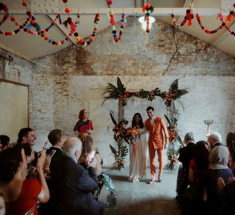 Industrial wedding ceremony with colourful pompom ceiling decorations and a tropical flower altar 