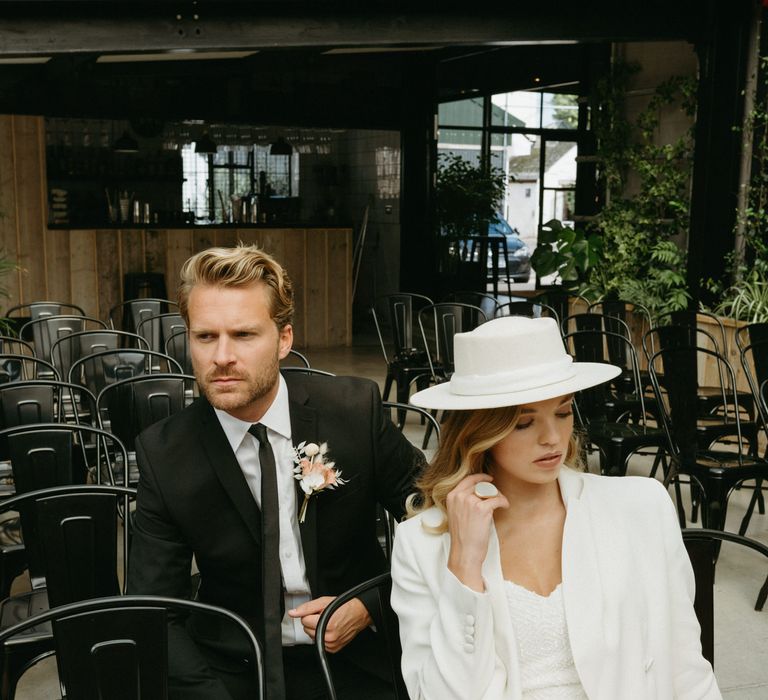 Stylish bride in a sequin jumpsuit, blazer and bridal hat sitting on the black metal chairs with her groom at The Shack Revolution wedding venue 