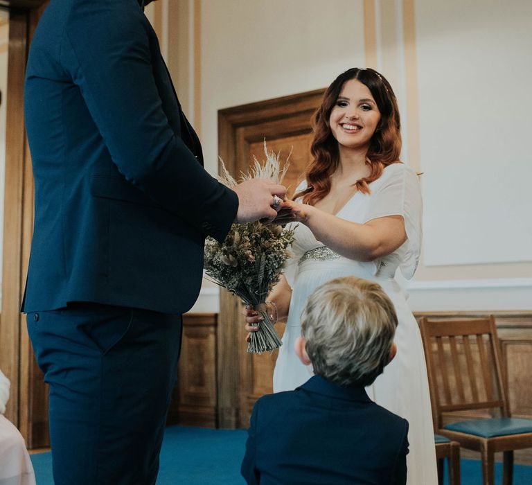 Groom in navy suit holding hands with smiling bride holding dried flower bouquet