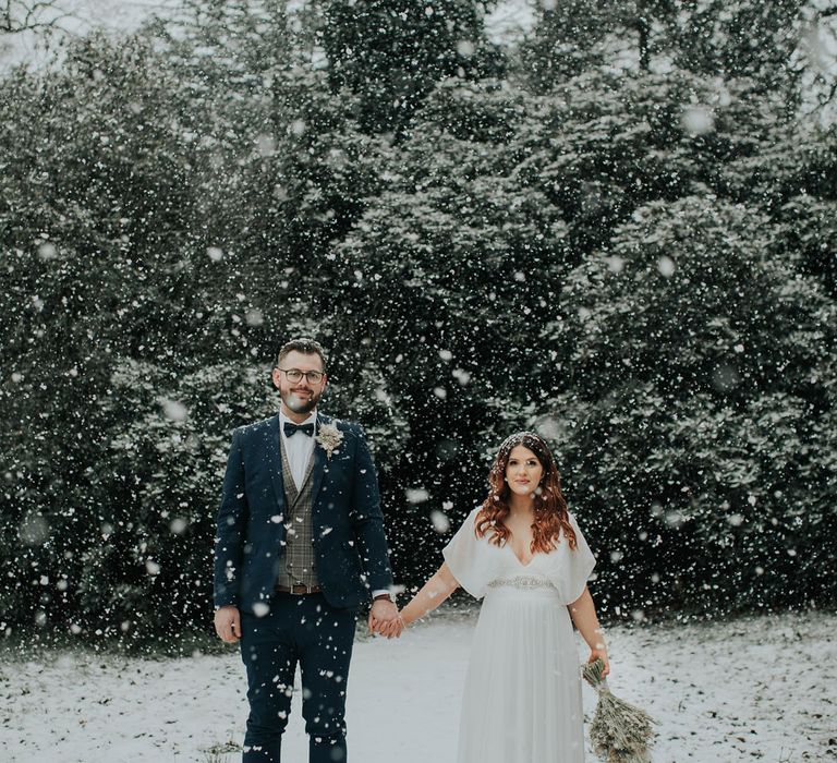 Bride in white wedding dress with silver detailing holding dried flower bouquet holds hands with groom in navy suit and tartan waistcoat during snowfall at Cannon Hall