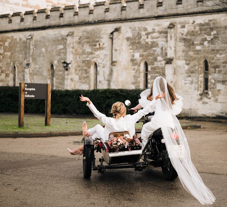 Bride riding a motorcycle with her cathedral length veil trailing behind and her bride sitting in a side car in a jacket waving their hands in the air 