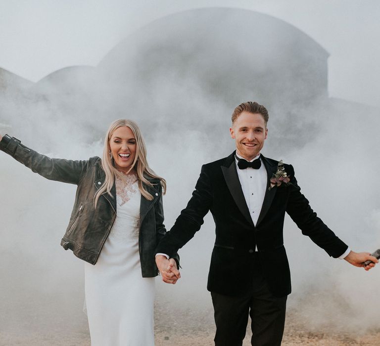 Bride in personalised leather bridal jacket and white Charlie Brear wedding dress holding hands with groom in black Hugo Boss suit holding white smoke bombs