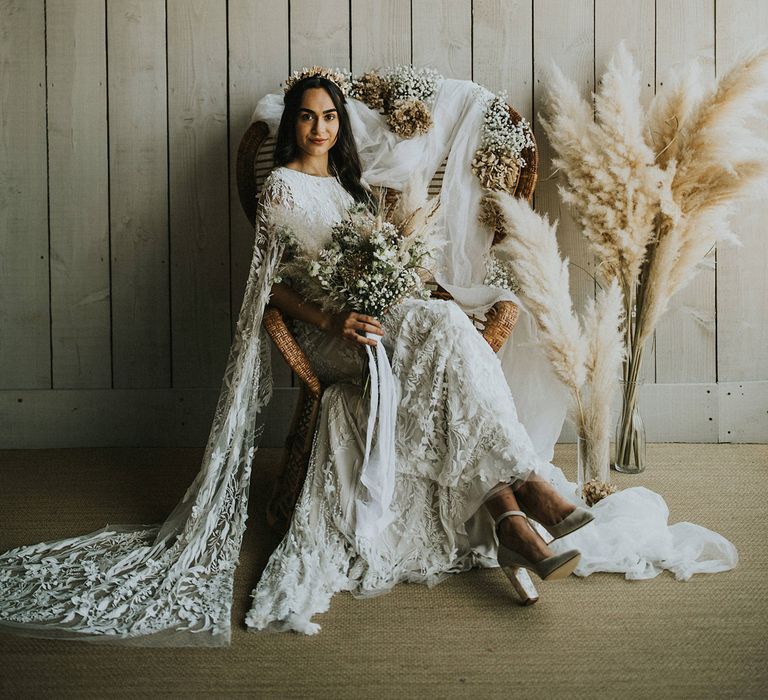 Boho bride in a lace wedding dress holding a dried flower bouquet sitting on a wicker chair 