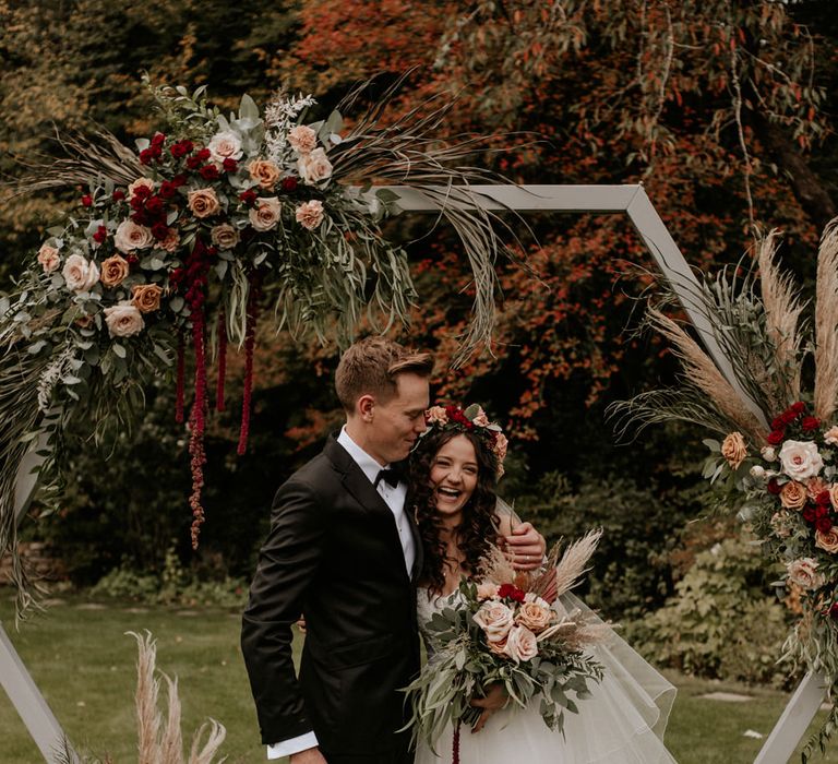 Groom in a tuxedo and bride in a tulle skirt standing in front of a wooden frame decorated with red and green flowers 