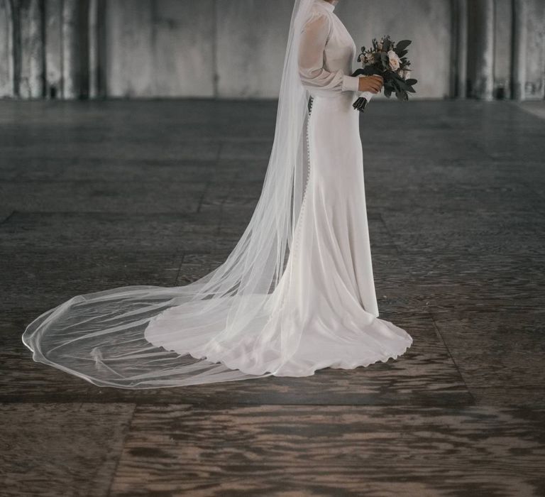 Bride stands in grey room whilst holding floral bouquet as veil and gown is spread out around her