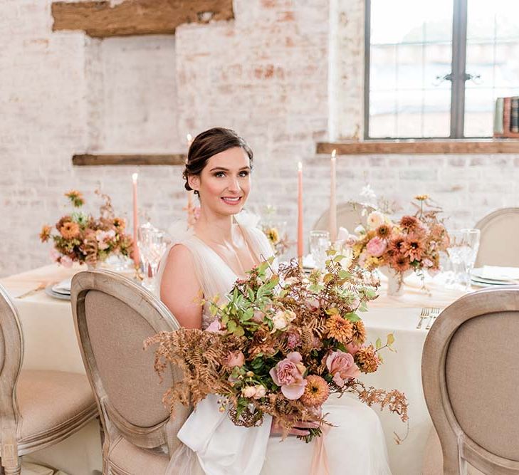 Bride in a tulle wedding dress sitting at Dorfold hall reception table holding an autumn bouquet with complementing centrepiece flowers 